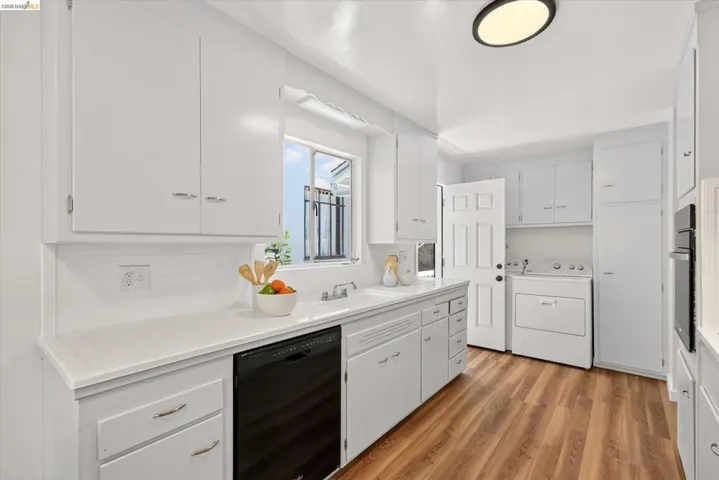 Kitchen featuring extensive white cabinetry, light countertops, wood-finish flooring, a built-in dishwasher, and a window above the sink