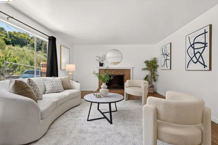 Spacious living area featuring a brick fireplace, large picture window, white walls, wood-finish flooring, and a light-toned area rug