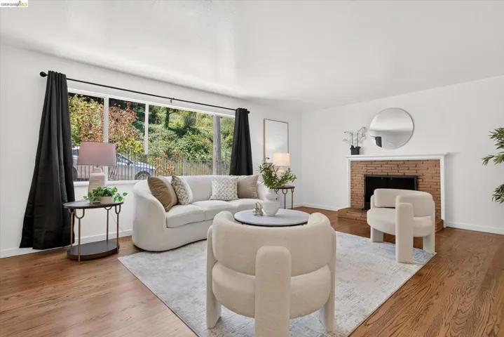 Spacious living area featuring expansive windows, wood-finish flooring, a brick fireplace with a white mantel, and bright white walls
