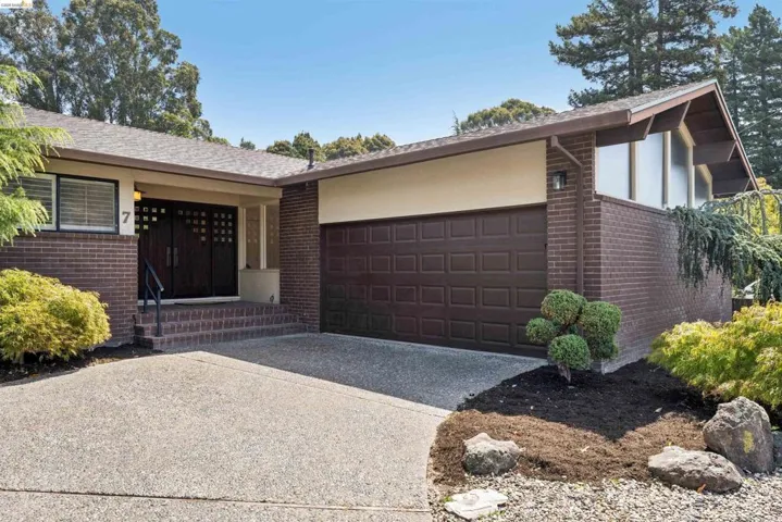 Brick facade residence featuring a two-car garage, mature landscaping, and an exposed aggregate driveway