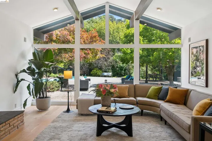 Vaulted ceiling living area featuring a large picture window, exposed structural beams, light wood-finish flooring, and a brick hearth