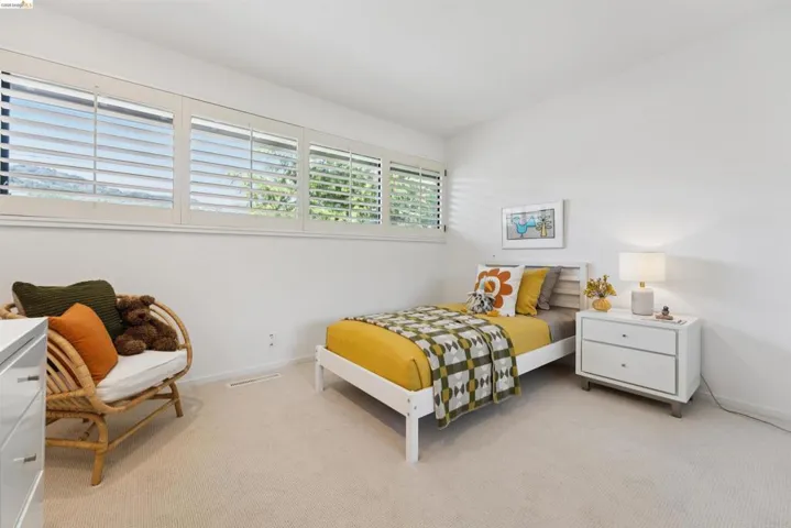 Bright bedroom featuring multiple windows with plantation shutters, neutral carpeting, and a built-in wall-mounted air vent