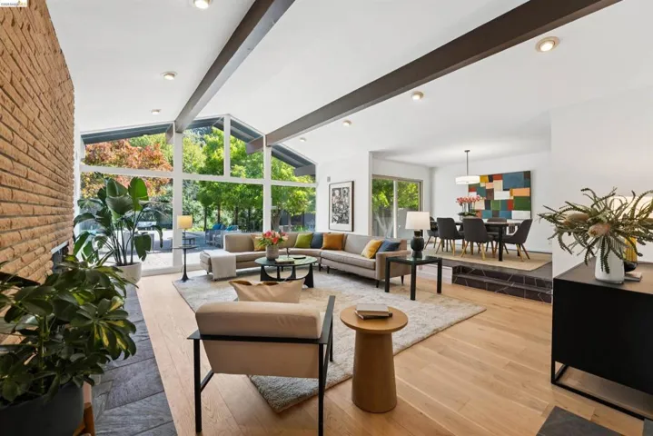 Open-concept living area featuring a brick fireplace with a slate hearth, vaulted ceilings with exposed beams, and expansive floor-to-ceiling windows