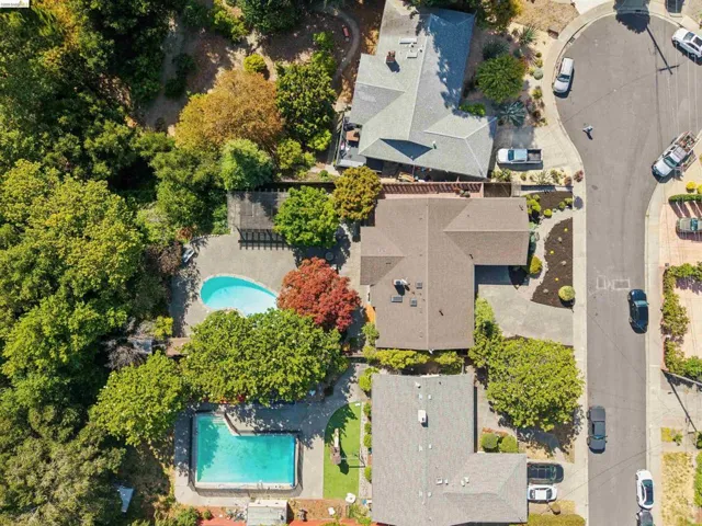 Aerial perspective showcasing a property with a distinct L-shaped roofline, mature surrounding trees, a paved driveway, and established landscaping