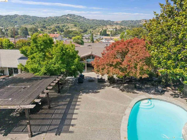 Expansive outdoor area featuring a freeform swimming pool, an exposed aggregate patio, a large pergola, and mature trees providing natural shade