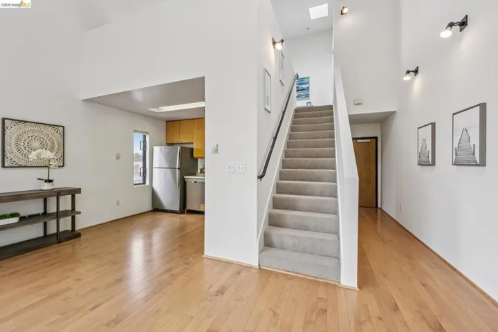 Open-concept living area featuring light wood-finish flooring, a carpeted staircase with a black handrail, a skylight, and a partial view of a kitchen with light wood cabinetry and stainless steel appliances