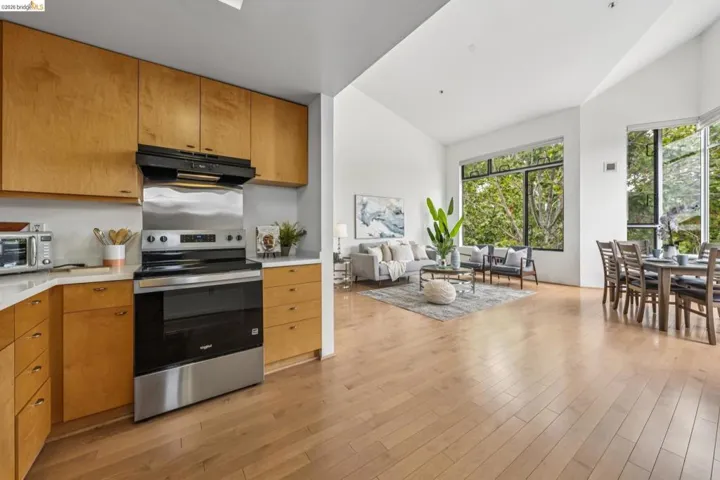 Open-concept living space featuring wood-finish flooring, high ceilings, large windows, a kitchen with light wood cabinetry, and stainless steel appliances