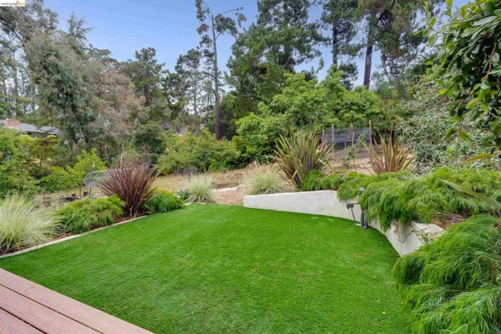 Decking overlooking a landscaped yard featuring artificial turf, mature trees, a retaining wall, and integrated outdoor lighting