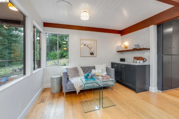 Bright interior space featuring a window wall, wood-finish flooring, exposed ceiling beams, and a built-in wet bar with dark cabinetry, a stone countertop, and an open wood shelf