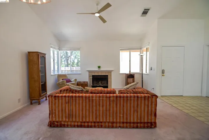 Living room featuring a vaulted ceiling with a ceiling fan, a fireplace with a tiled surround, multiple windows, and a combination of carpet and tiled flooring