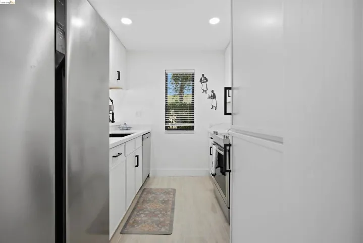 Galley kitchen featuring stainless steel appliances, white cabinetry with black hardware, recessed lighting, and light wood-finish flooring