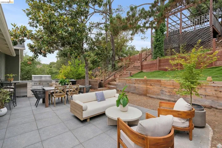 Expansive outdoor patio featuring large format pavers, a built-in BBQ station with stone surround, a multi-tiered backyard with wood retaining walls, and an elevated wood deck