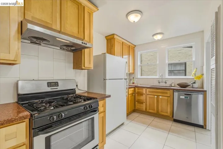 Kitchen featuring natural wood cabinetry, white tile flooring, dual windows, stainless steel dishwasher, and a built-in range with an overhead exhaust hood
