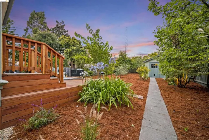 Expansive backyard featuring a wood deck with railings, a stone pathway, landscaping with mulch and established plantings, and a detached shed
