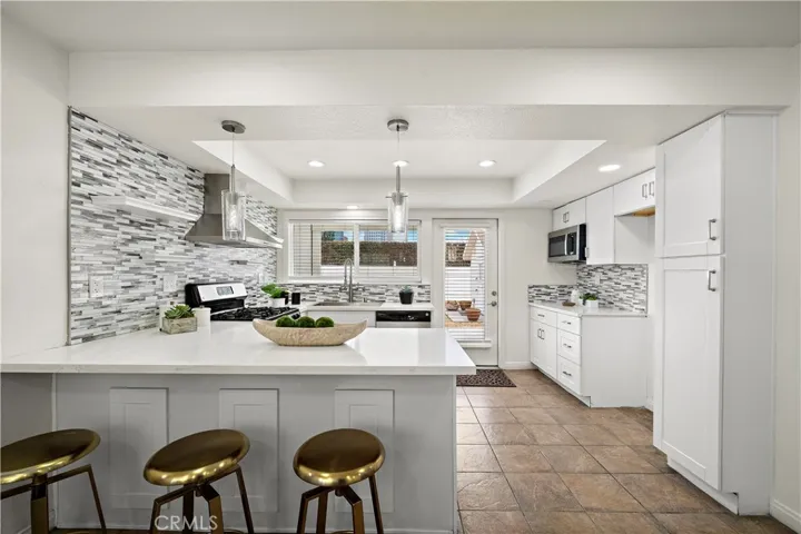 Breakfast bar with pendant lighting, quartz countertops, glass mosaic tile backsplash, floating open shelving, stainless appliances (gas range, vent hood, dishwasher, and built-in microwave).