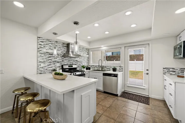 Quartz countertops, glass mosaic tile backsplash, floating open shelving, breakfast bar with chic pendant lighting, deep stainless sink with designer faucet, and stainless steel appliances including gas range, vent hood, dishwasher, and built-in microwave.
