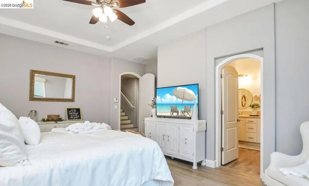 Bedroom with light wood-style flooring, a ceiling fan, a raised ceiling, connected bathroom, and arched walkways