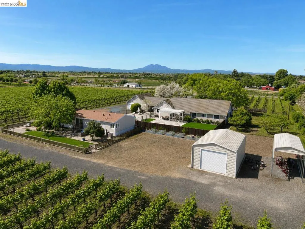 Expansive agricultural property featuring established vineyards, a gravel driveway, a detached shed with a roll-up door, a separate covered parking structure, and a large dwelling with an attached covered patio