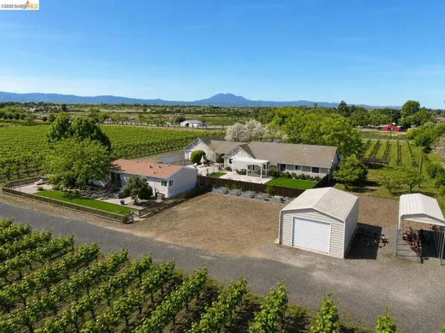Expansive agricultural property featuring established vineyards, a gravel driveway, a detached shed with a roll-up door, a separate covered parking structure, and a large dwelling with an attached covered patio