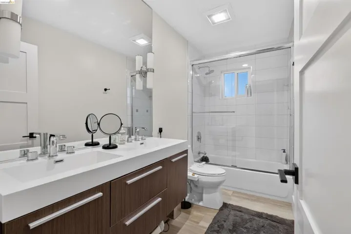 Modern bathroom featuring a dual vanity with wood-finish cabinetry, a white integrated countertop with two sinks, a tub-shower combination with a glass enclosure and white subway tile surround, and light wood-look flooring