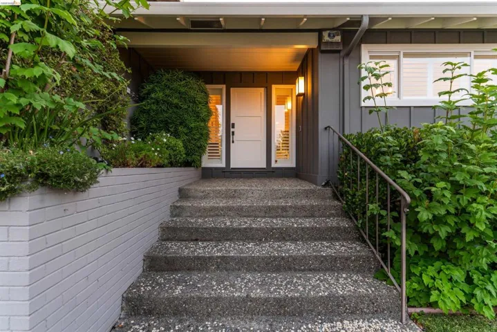 Entryway featuring exposed aggregate steps with a metal handrail, a painted brick retaining wall, and verdant landscaping