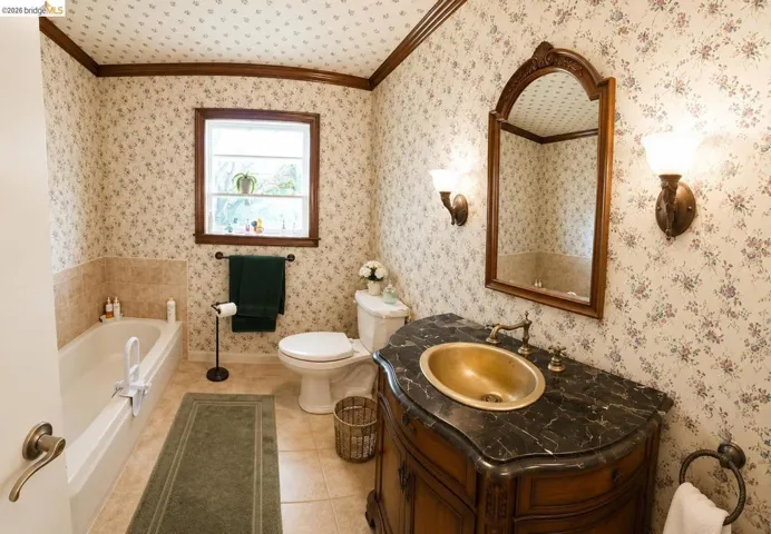 Bathroom featuring a built-in tub with beige tiled surround, a window with a wood frame, and a vanity with a dark countertop and brass basin