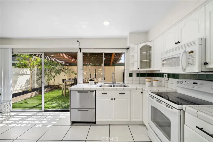 Bright white kitchen with tile flooring, tile countertops, white shaker cabinetry, peninsula with bar seating, gas range, built-in microwave, and Fisher & Paykel two-drawer dishwasher.