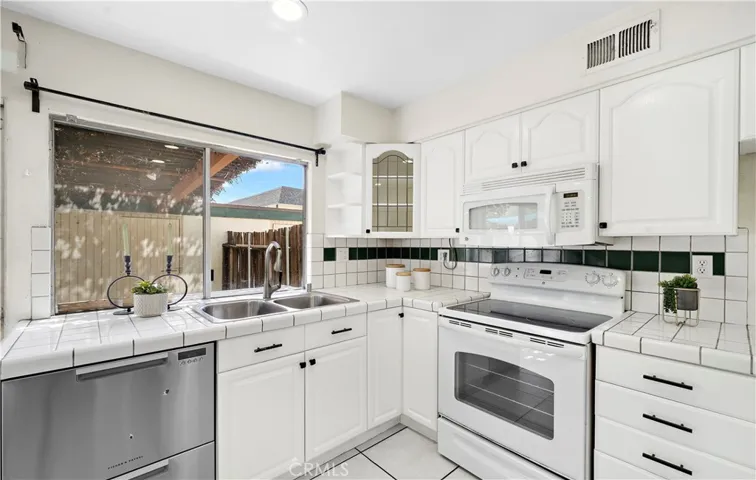 Large light and bright white kitchen with tile flooring, tile countertops, white shaker cabinetry, peninsula with bar seating, gas range, built-in microwave, and Fisher & Paykel two-drawer dishwasher.