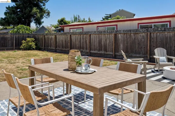Concrete patio featuring a slatted outdoor dining table, a patterned area rug, and a separate seating area with Adirondack chairs and a fire pit