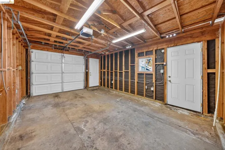 Single-car garage featuring a paneled white garage door, concrete flooring, exposed wood framing, and fluorescent overhead lighting