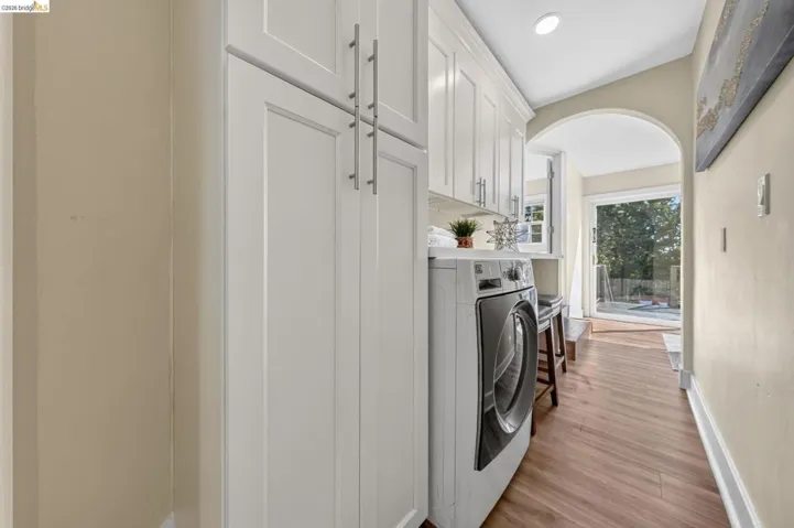 Laundry area with white shaker cabinetry, stainless steel front-load appliance, wood-finish flooring, and an arched doorway opening to exterior access