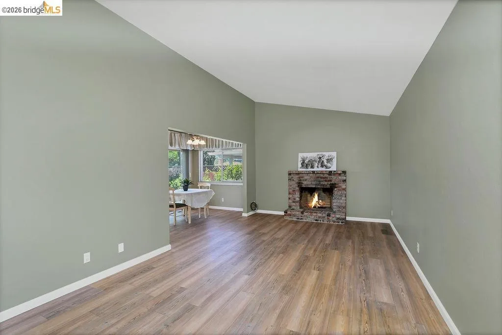 Living area featuring a brick fireplace, wood-finish flooring, vaulted ceilings, and a framed opening to an additional room with multiple windows