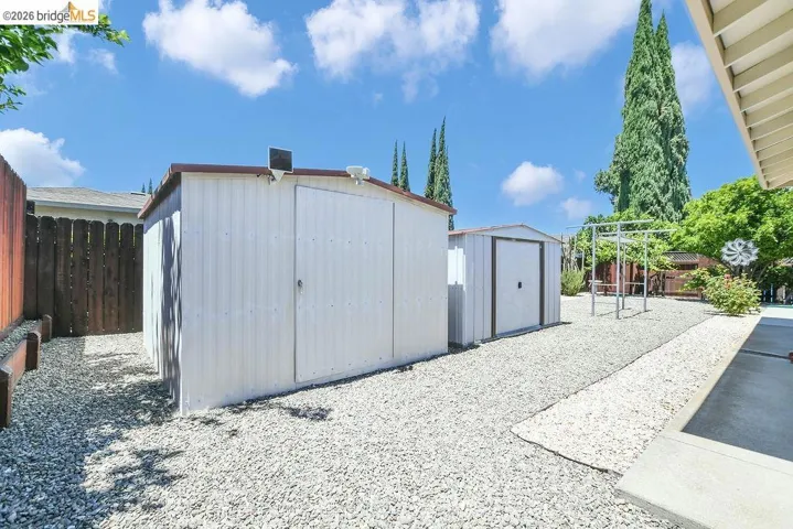 Gravel-covered outdoor space featuring two white storage sheds with brown trim, a wooden fence, and mature trees
