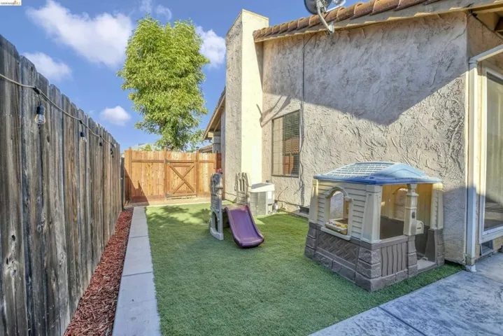 Private side yard featuring artificial turf, a wood fence with string lighting, a concrete pathway, and a stucco exterior wall