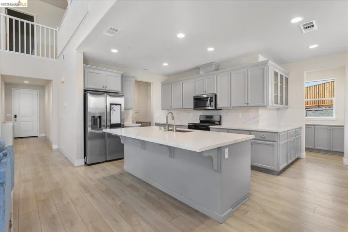 Open-concept kitchen featuring an oversized island with countertop seating, light gray cabinetry, stainless steel appliances, and wood-finish flooring