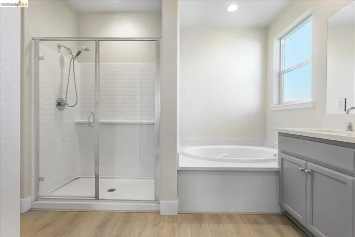Contemporary bathroom featuring a glass-enclosed shower with subway tile, a built-in soaking tub, a double vanity with gray cabinetry, a window, and wood-finish flooring