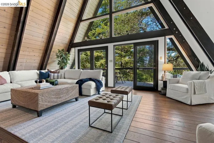 Spacious living area featuring expansive A-frame windows, wood-finish flooring, exposed architectural beams, and a double glass door balcony entrance