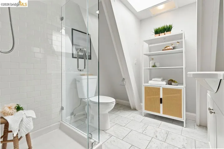 Glass-enclosed shower with white subway tile, white toilet, skylight, light-colored tile flooring, and a white storage unit with woven cane doors