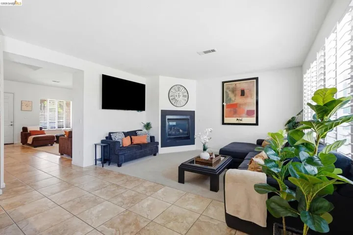 Open-concept living space featuring a corner fireplace with dark surround, light-toned tile flooring, and large windows with plantation shutters