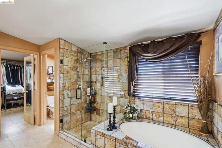 Stone-tiled bathroom featuring a glass-enclosed shower, built-in soaking tub, and natural wood blinds