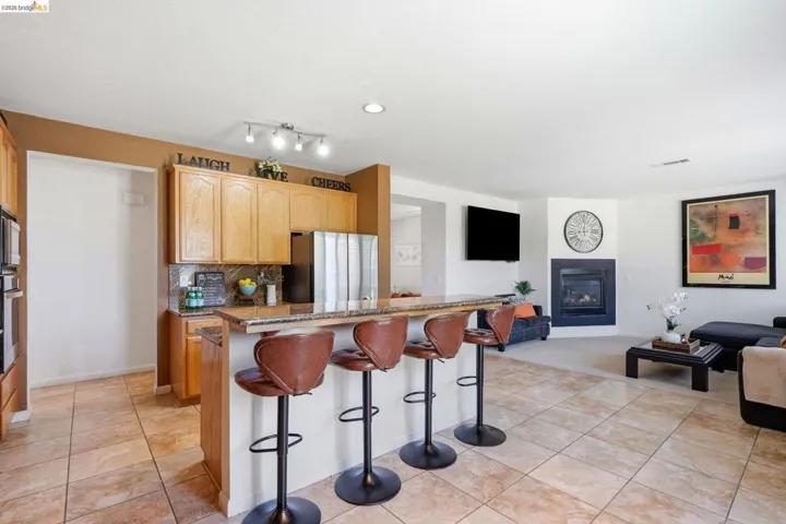 Open-concept living space featuring a breakfast bar, stone-finish countertops, warm wood cabinetry, stainless steel appliances, and a corner fireplace with a dark surround