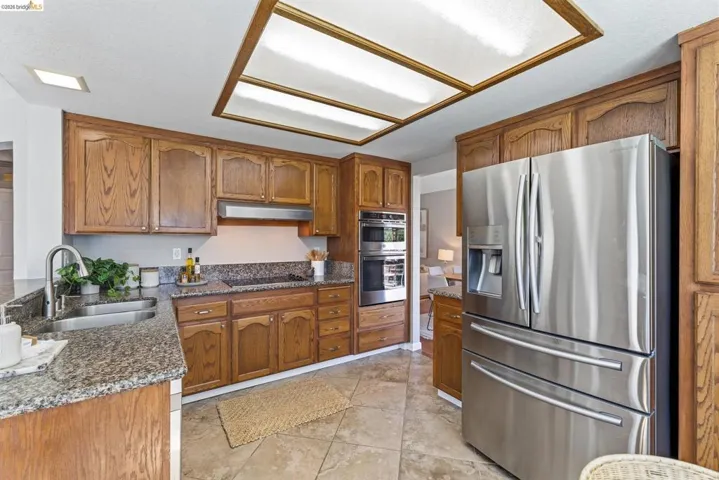 Kitchen featuring wood cabinetry, stone countertops, a stainless steel double basin sink, a built-in double oven, and a stainless steel refrigerator