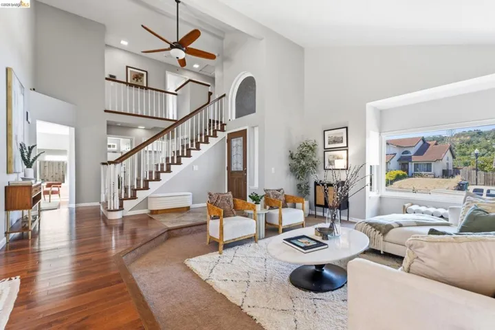 Foyer and living area featuring high ceilings with a ceiling fan, a grand staircase with wood-finish handrails and white balusters, wood-finish flooring, and a large bay window