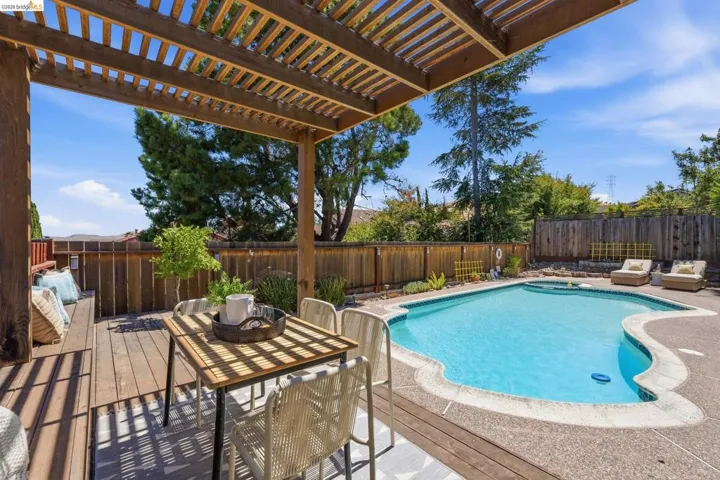 Wood deck with pergola covering a dining area, overlooking a pool with a concrete surround and a wood fence