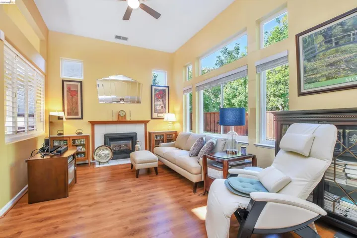 Vaulted ceiling living area featuring wood-finish flooring, a fireplace with a tile surround and wood mantel, multiple windows with plantation shutters, transom windows, and a ceiling fan