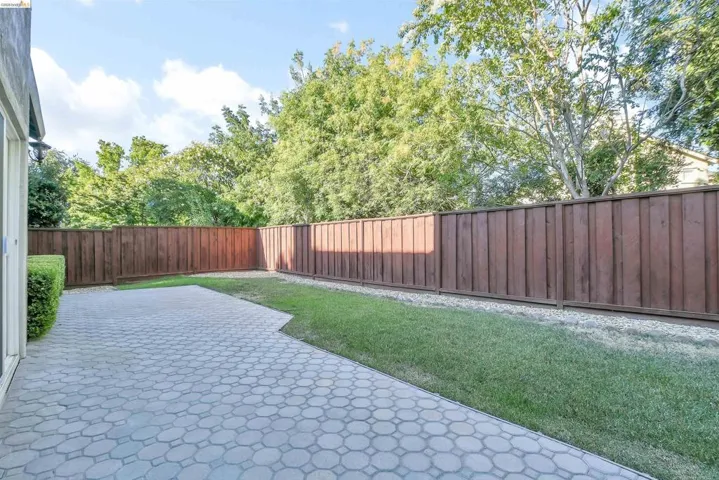 Outdoor patio featuring geometric pavers, perimeter wooden fencing, a grass area, and mature trees