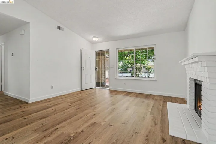 Spacious living area featuring wood-finish flooring, a white brick fireplace, and a large window with security bars