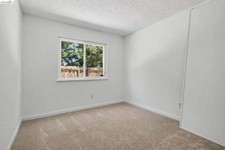 Bright room featuring a window with white trim, tan carpeting, white walls, and a popcorn ceiling