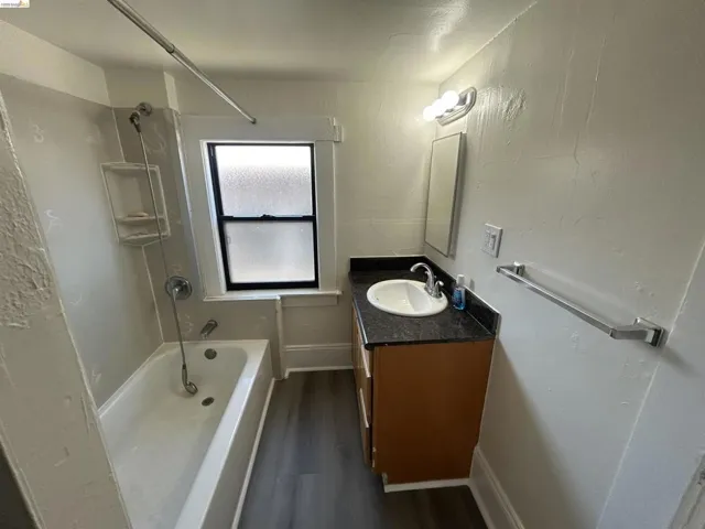 Bathroom featuring a built-in bathtub with shower, a vanity with a dark countertop and integrated sink, wood-finish flooring, a wall-mounted mirror, and a frosted window
