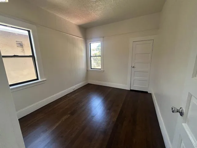 Room featuring dark wood-finish flooring, two window openings, white painted walls, and a white paneled door with a silver knob